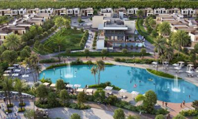 Aerial view of Sobha Realty’s luxury villa community showing the central clubhouse overlooking a landscaped lagoon with fountains, people walking along pathways, sun loungers by the water, palm trees, and modern villas arranged in the background.