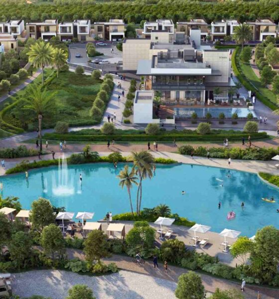 Aerial view of Sobha Realty’s luxury villa community showing the central clubhouse overlooking a landscaped lagoon with fountains, people walking along pathways, sun loungers by the water, palm trees, and modern villas arranged in the background.