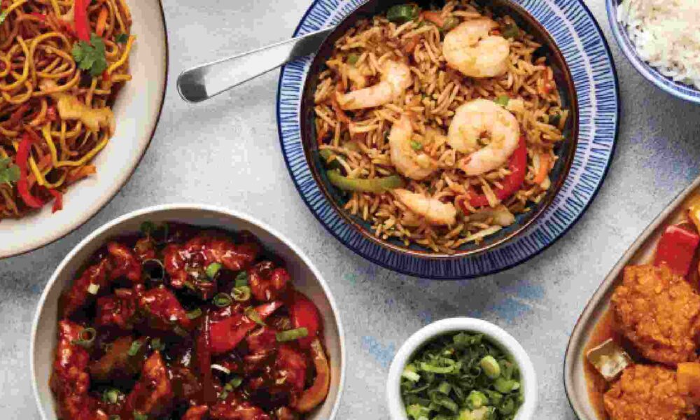 Overhead view of assorted Chinese dishes from IKEA Restaurant, including stir-fried noodles, fried rice topped with prawns, sweet and sour chicken, crispy chicken in sauce, and a bowl of steamed white rice arranged on a light table.