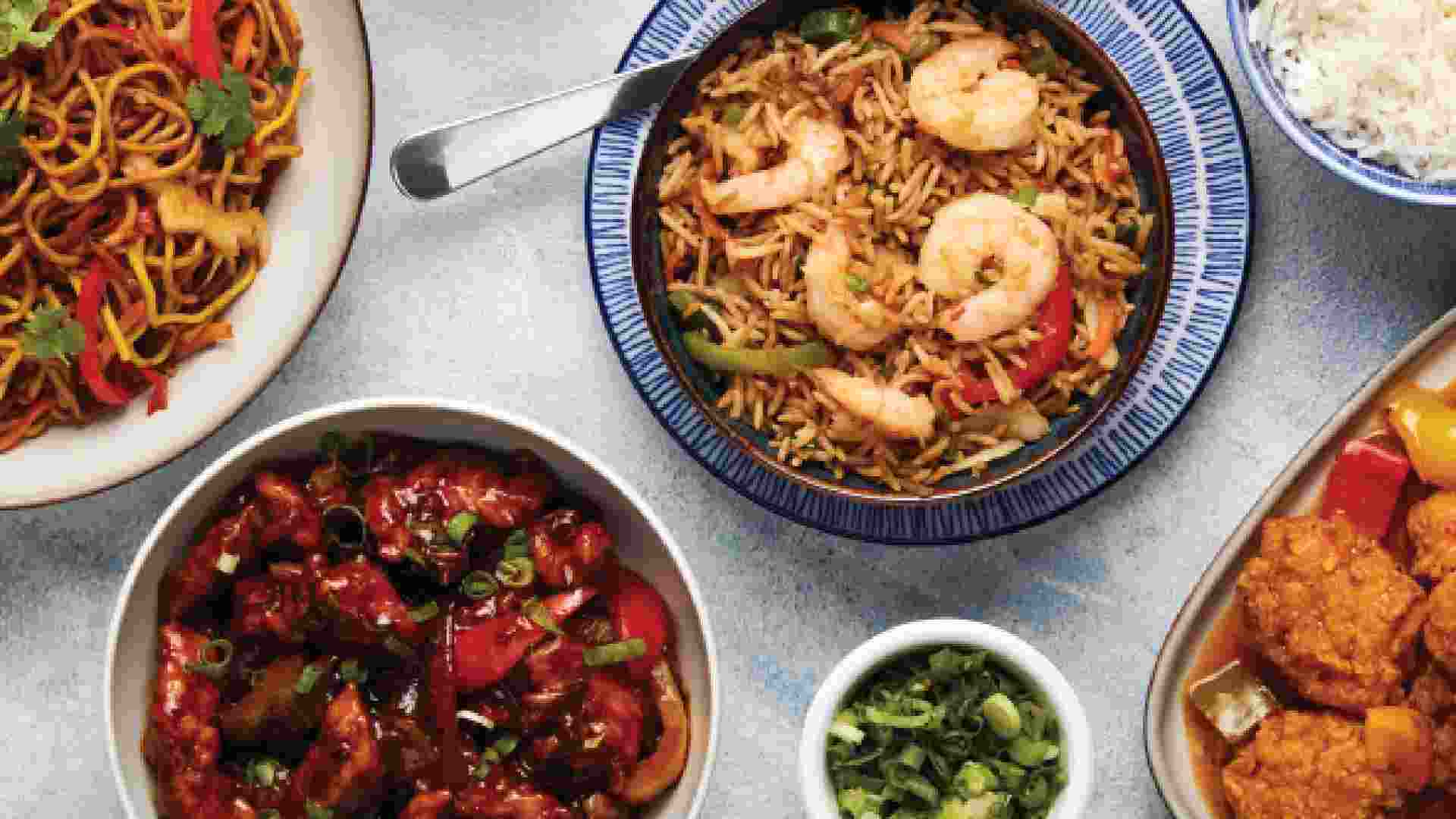 Overhead view of assorted Chinese dishes from IKEA Restaurant, including stir-fried noodles, fried rice topped with prawns, sweet and sour chicken, crispy chicken in sauce, and a bowl of steamed white rice arranged on a light table.