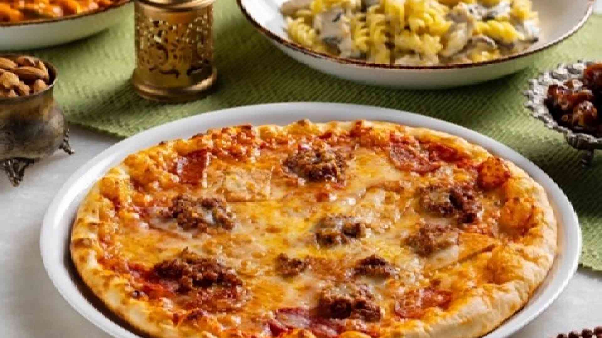 Table spread of dishes at The Big Chill Café featuring a cheese pizza in the foreground, plates of creamy pasta, saffron rice, stir‑fried noodles, and flatbread, arranged with decorative lanterns in a warm café setting
