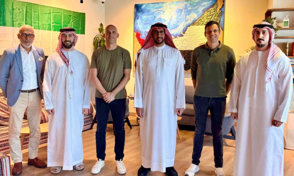 Group of six men standing indoors in a shared lounge-style setting, wearing a mix of traditional Emirati attire and smart-casual clothing, positioned side by side in front of sofas, patterned cushions, shelves with books and decor, and large colorful wall artworks, at a SaladStop-related meeting or partnership gathering.