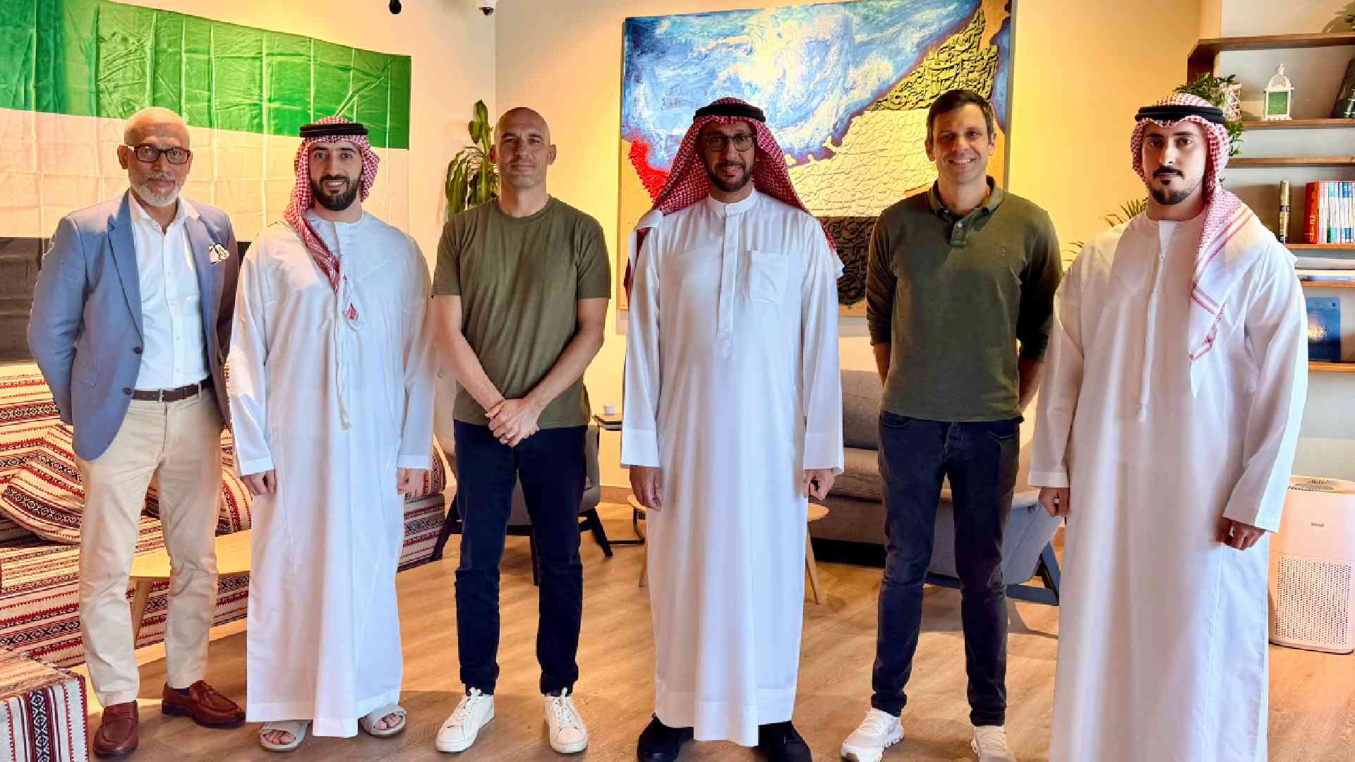 Group of six men standing indoors in a shared lounge-style setting, wearing a mix of traditional Emirati attire and smart-casual clothing, positioned side by side in front of sofas, patterned cushions, shelves with books and decor, and large colorful wall artworks, at a SaladStop-related meeting or partnership gathering.