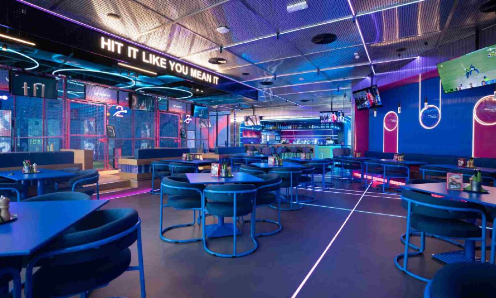 Interior of WIKIT at Emirates Golf Club showing neon‑lit AR cricket batting lanes, blue dining tables and chairs, digital screens, and a bar area with the slogan “Hit It Like You Mean It” displayed above.