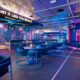 Interior of WIKIT at Emirates Golf Club showing neon‑lit AR cricket batting lanes, blue dining tables and chairs, digital screens, and a bar area with the slogan “Hit It Like You Mean It” displayed above.