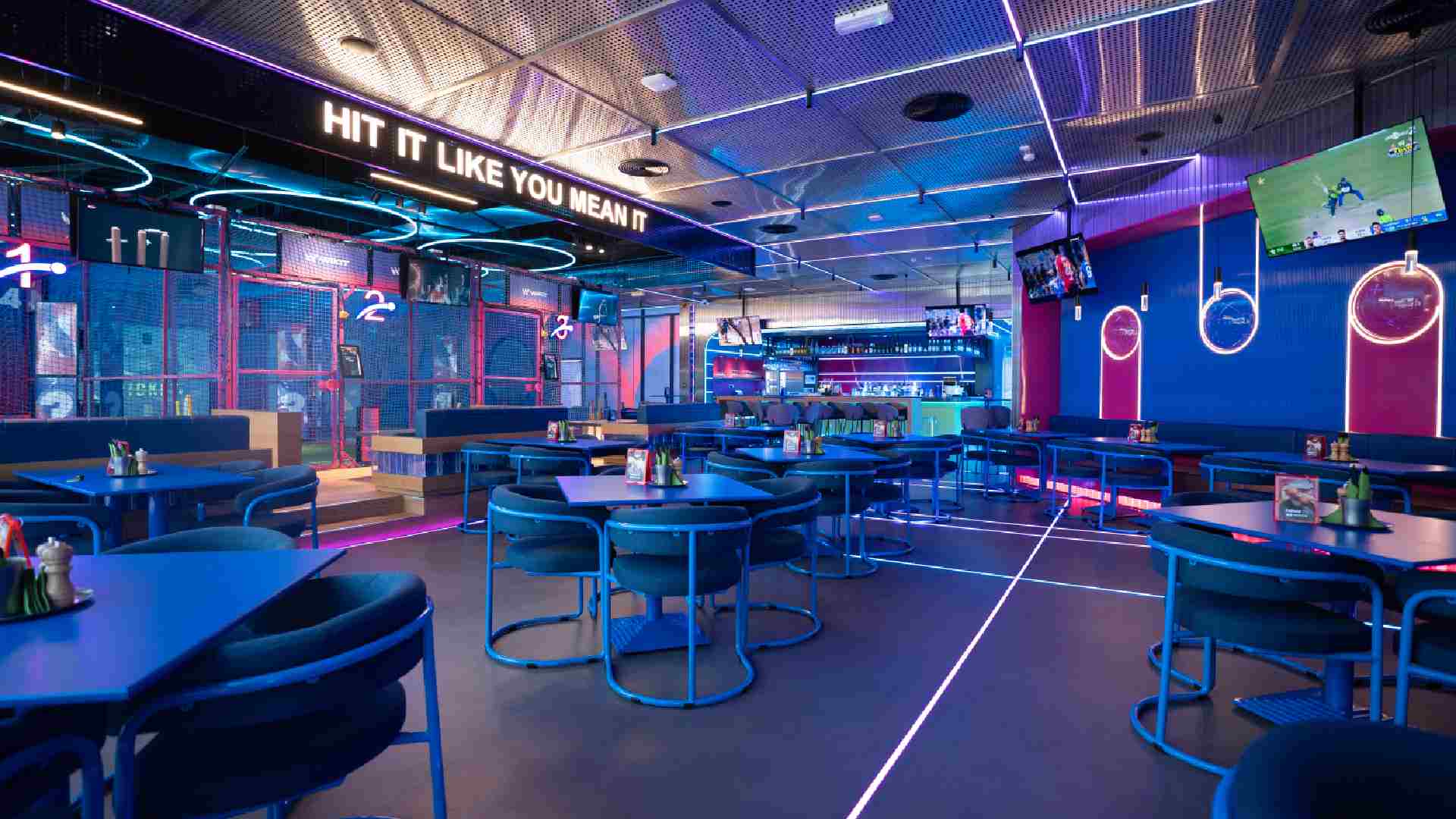 Interior of WIKIT at Emirates Golf Club showing neon‑lit AR cricket batting lanes, blue dining tables and chairs, digital screens, and a bar area with the slogan “Hit It Like You Mean It” displayed above.