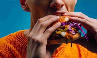 Close-up of a person biting into a burger made with Kerry ingredients, featuring a sesame bun, beef patty, melted cheese, jalapeños, purple cabbage slaw and dripping sauce against a blue background.