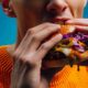 Close-up of a person biting into a burger made with Kerry ingredients, featuring a sesame bun, beef patty, melted cheese, jalapeños, purple cabbage slaw and dripping sauce against a blue background.