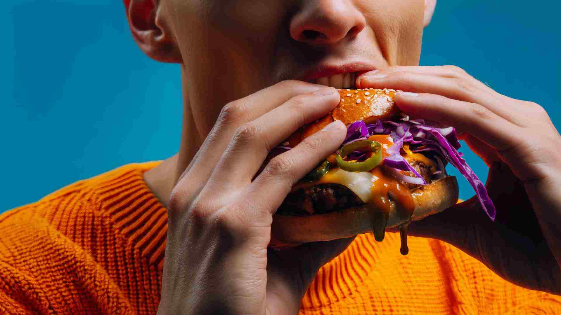 Close-up of a person biting into a burger made with Kerry ingredients, featuring a sesame bun, beef patty, melted cheese, jalapeños, purple cabbage slaw and dripping sauce against a blue background.