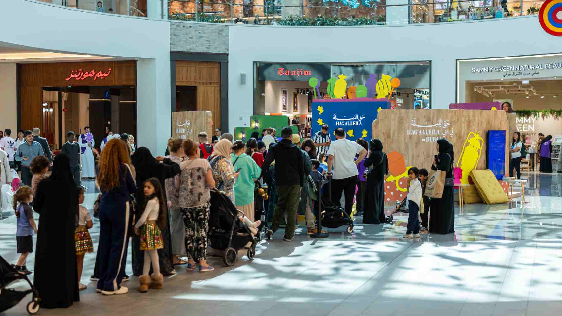 Families and children queue around a colourful HAG AL LEILA activation inside a Dubai shopping mall, where staff distribute sweets and lead festive activities in a decorated central atrium