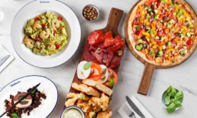 Overhead view of Carluccio’s Italian spread featuring mixed vegetable pizza on a wooden board, antipasti platter with cured meats, mozzarella, bread and dips, avocado pasta, and a plated dessert on a marble table