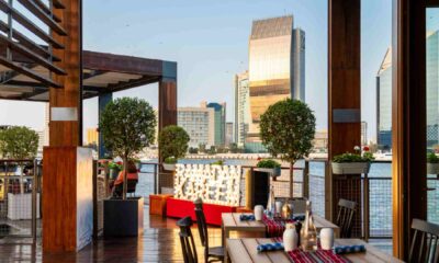 Outdoor dining area at Skafos overlooking Dubai Creek, featuring wooden tables set for guests, decorative greenery, warm lighting, and a clear view of modern waterfront buildings at sunset.