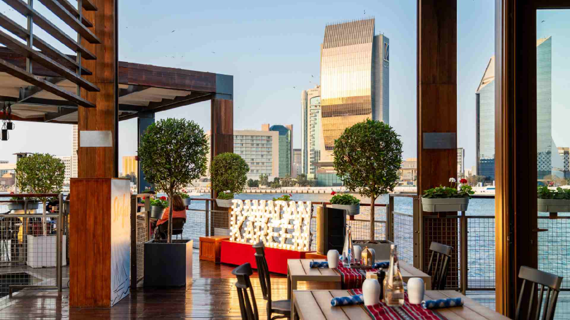 Outdoor dining area at Skafos overlooking Dubai Creek, featuring wooden tables set for guests, decorative greenery, warm lighting, and a clear view of modern waterfront buildings at sunset.