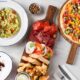 Overhead view of Carluccio’s Italian spread featuring mixed vegetable pizza on a wooden board, antipasti platter with cured meats, mozzarella, bread and dips, avocado pasta, and a plated dessert on a marble table