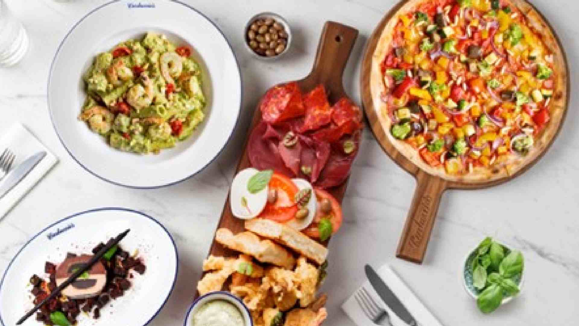 Overhead view of Carluccio’s Italian spread featuring mixed vegetable pizza on a wooden board, antipasti platter with cured meats, mozzarella, bread and dips, avocado pasta, and a plated dessert on a marble table