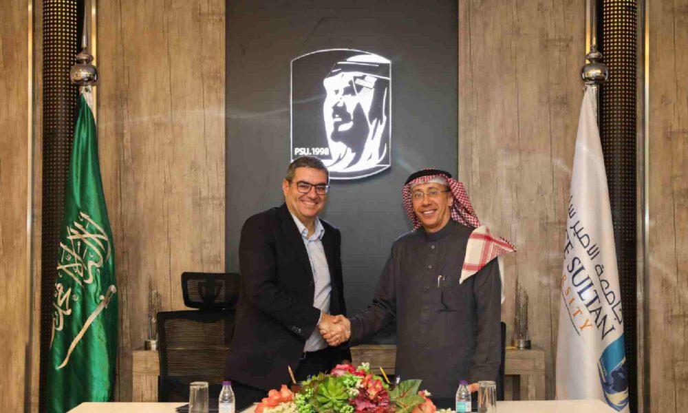 Two individuals shaking hands across a table during a formal signing event, with the Saudi Arabian flag on the left, the Prince Sultan University flag on the right, and the university’s emblem displayed on the wall behind them.