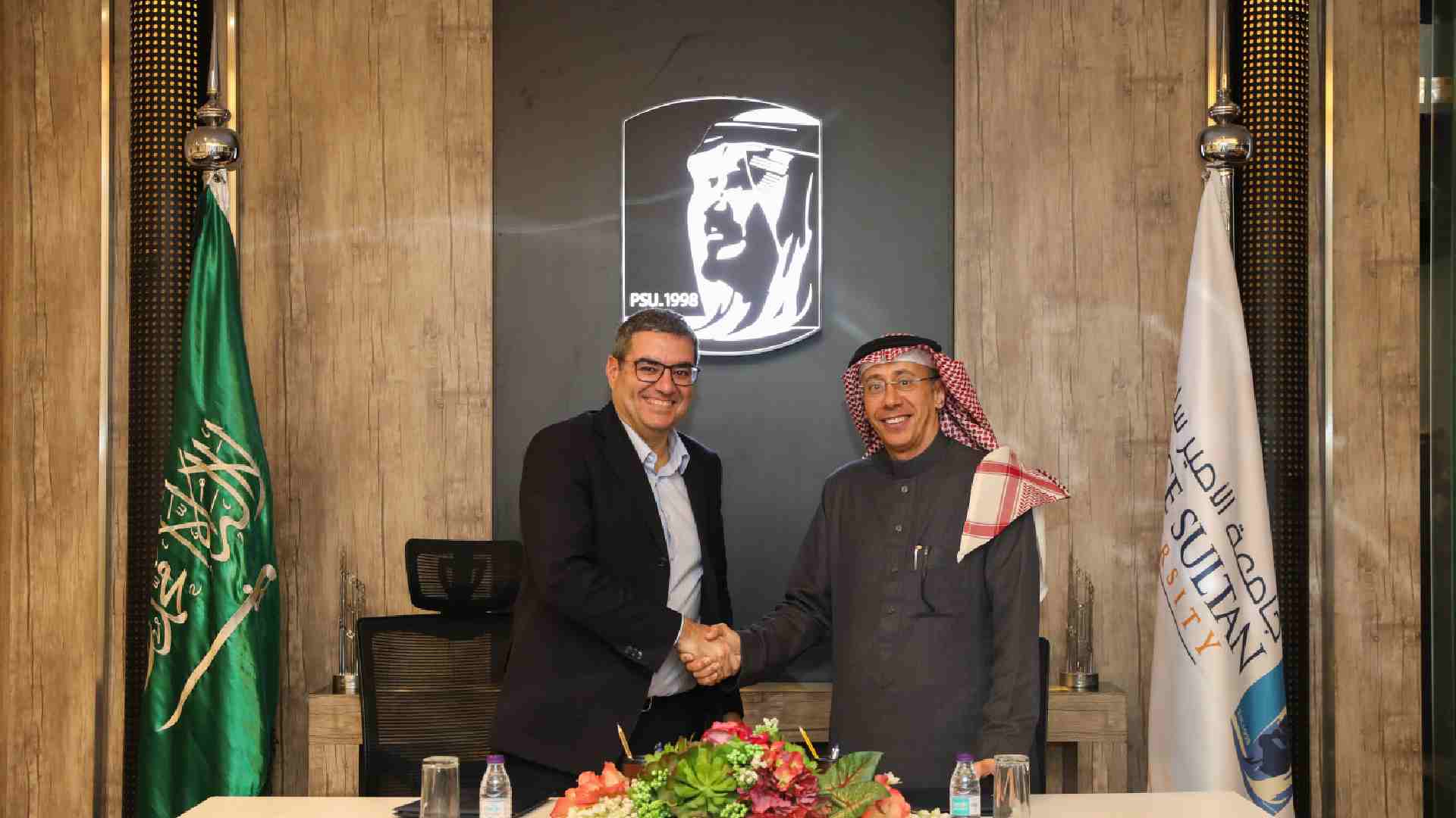 Two individuals shaking hands across a table during a formal signing event, with the Saudi Arabian flag on the left, the Prince Sultan University flag on the right, and the university’s emblem displayed on the wall behind them.