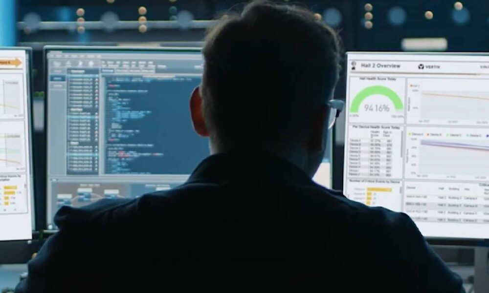 A person sits in front of multiple computer monitors displaying system dashboards, performance analytics, and code, monitoring data center operations inside a Vertiv facility.
