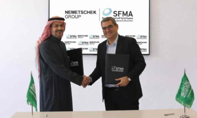 A representative from Nemetschek Group and a representative from the Saudi Facility Management Association shake hands while holding signed MoU folders, standing in front of a display showing both organizations’ logos, with Saudi flags placed on the table in front of them.