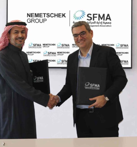 A representative from Nemetschek Group and a representative from the Saudi Facility Management Association shake hands while holding signed MoU folders, standing in front of a display showing both organizations’ logos, with Saudi flags placed on the table in front of them.