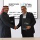 A representative from Nemetschek Group and a representative from the Saudi Facility Management Association shake hands while holding signed MoU folders, standing in front of a display showing both organizations’ logos, with Saudi flags placed on the table in front of them.