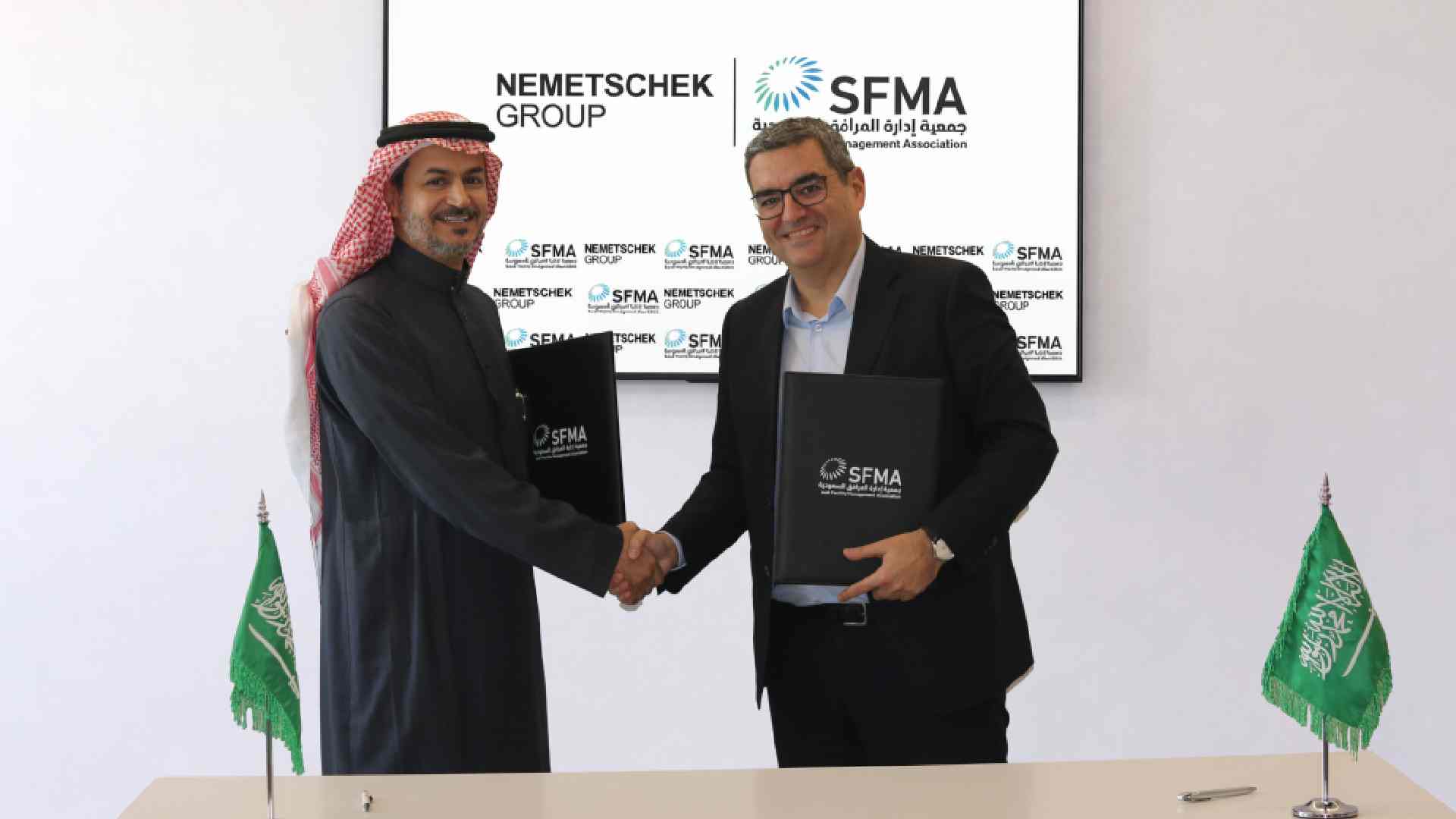 A representative from Nemetschek Group and a representative from the Saudi Facility Management Association shake hands while holding signed MoU folders, standing in front of a display showing both organizations’ logos, with Saudi flags placed on the table in front of them.