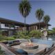 A modern luxury villa designed by NKEY Architects featuring a two‑storey contemporary structure with wood and glass facades, surrounded by palm trees, landscaped greenery, an infinity‑style swimming pool, a sculptural outdoor art piece, and a sunken seating area with a central fire pit in the foreground.