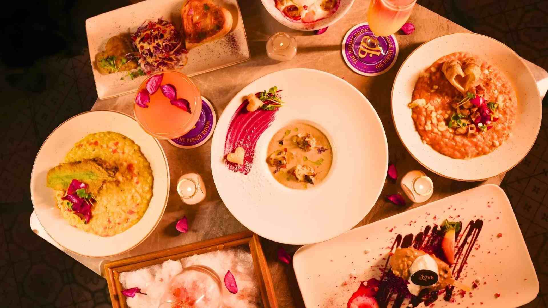 Overhead view of Valentine’s Week dishes and cocktails at THE PERMIT ROOM, featuring Indian mains, desserts with romantic plating, and pink-themed drinks on a dining table.