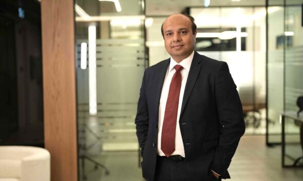 Executive from Dhruva Consultants standing in a modern office corridor, wearing a dark business suit and red tie, with glass meeting rooms and workspaces in the background.