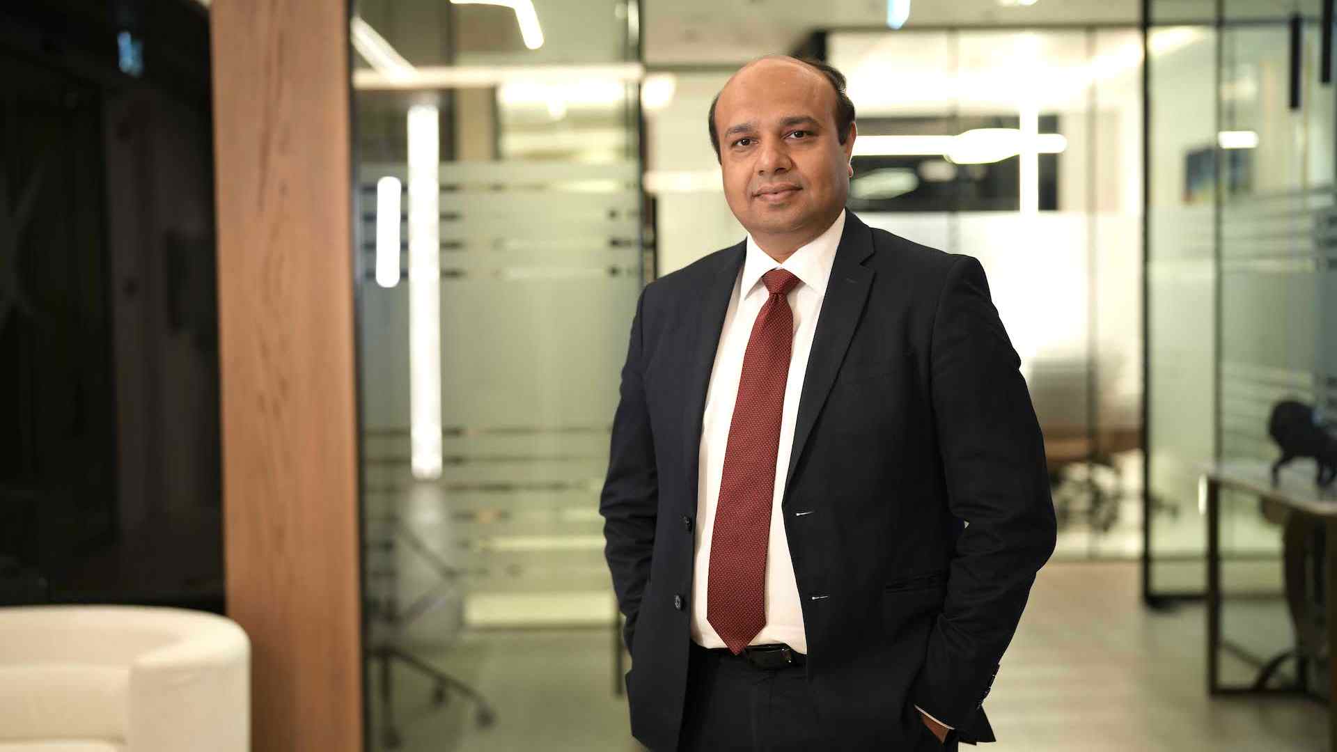 Executive from Dhruva Consultants standing in a modern office corridor, wearing a dark business suit and red tie, with glass meeting rooms and workspaces in the background.