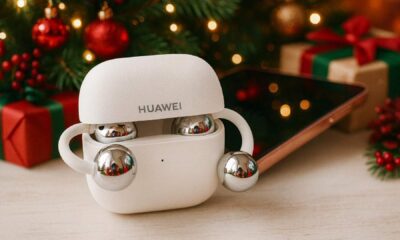 White HUAWEI FreeClip open‑ear earbuds inside an open charging case on a table, with a smartphone, Christmas tree, lights, and wrapped gift boxes in the background.