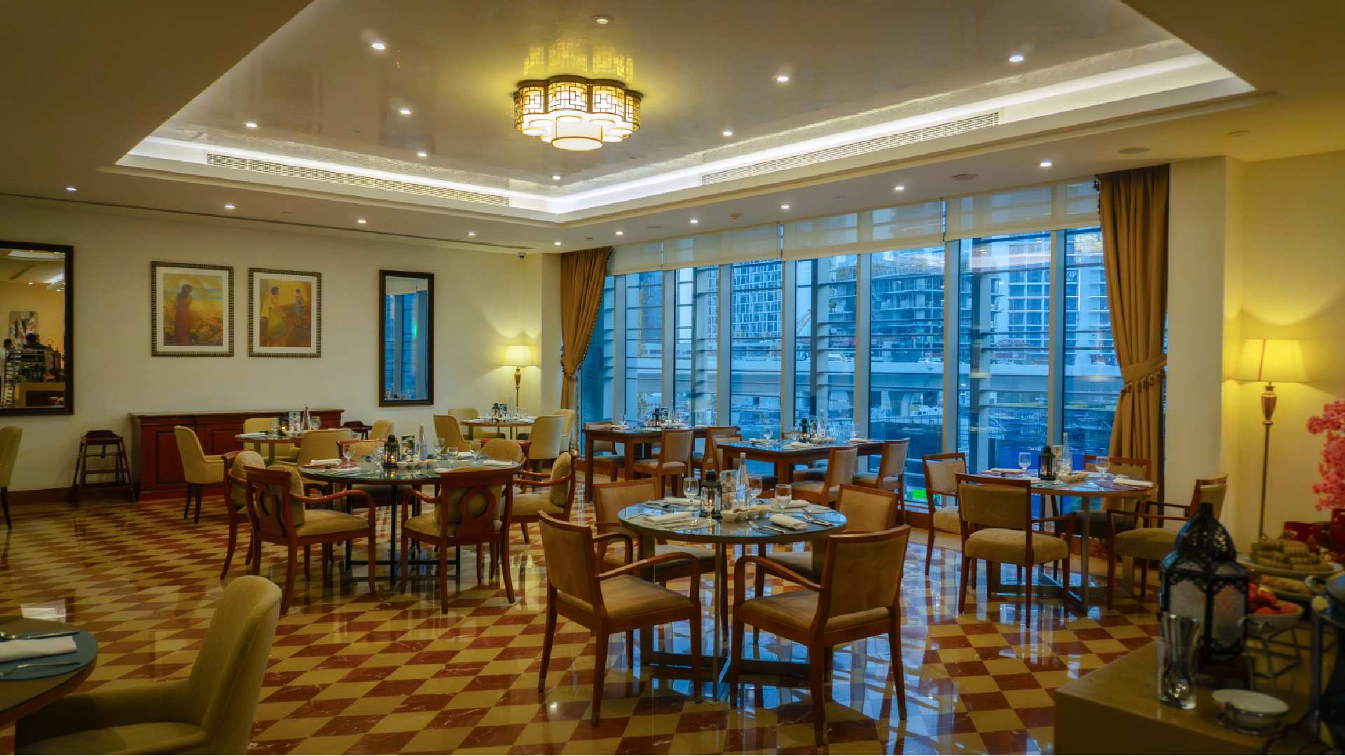 The image shows an elegant indoor dining space at Renaissance Business Bay Hotel. The room is arranged with multiple round and square wooden tables set with plates, glassware and cutlery, each surrounded by cushioned wooden chairs. The floor features a patterned tile design in warm tones, complementing the soft yellow lighting from ceiling fixtures and table lamps. Large floor‑to‑ceiling windows line one side of the room, offering views of nearby Dubai buildings and allowing natural light into the space. Framed artwork decorates the walls, and long curtains frame the windows, creating a refined and welcoming restaurant atmosphere.