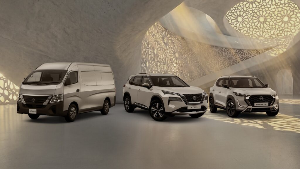 A wide shot of three white Nissan vehicles - the Urvan van, the X-Trail SUV, and the Magnite compact SUV - parked in a minimalist, sand-colored hall.