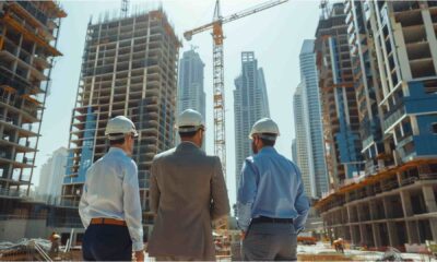 Three construction professionals wearing safety helmets stand on an active construction site in Dubai, facing multiple high-rise buildings under construction with tower cranes and concrete structures in progress, illustrating large-scale urban development and project execution.
